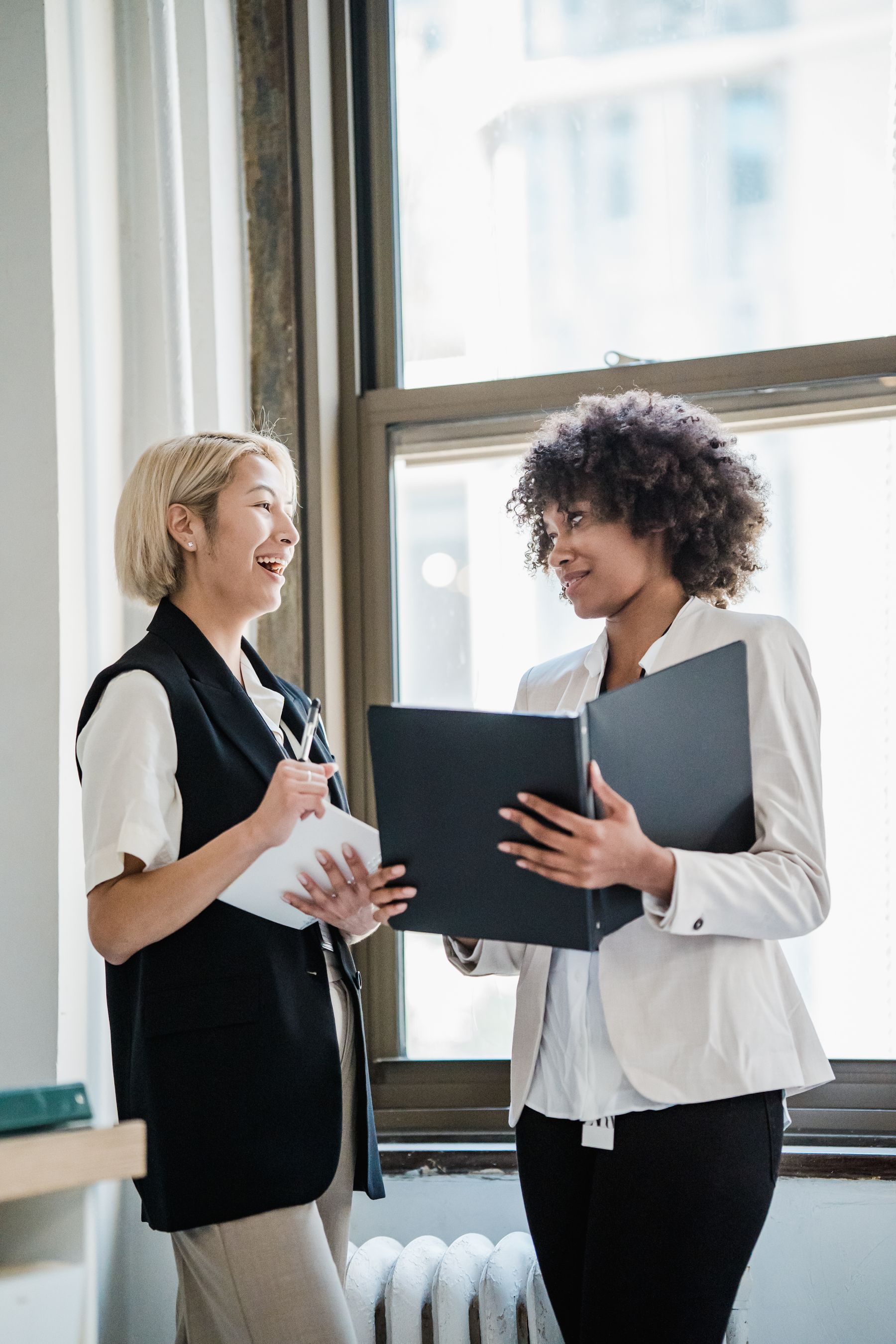 Two consultants smiling while reviewing a client folder near a bright office window